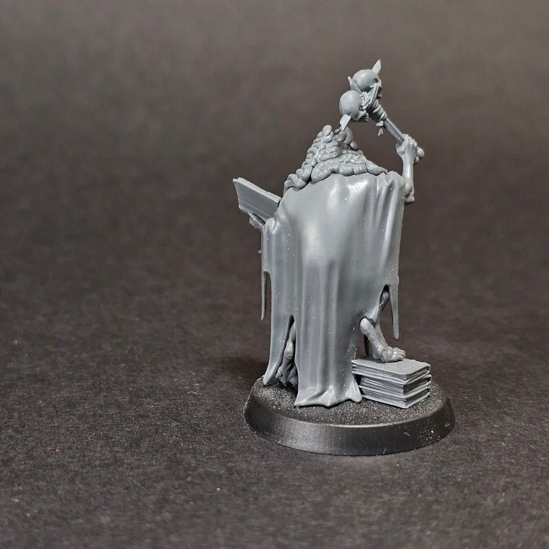 A used Warhammer Age of Sigmar: Flesh Eater Courts Grand Justice Gormayne miniature stands ghostly in gray, draped in tattered cloth and holding a staff atop a round base with books, set against a dark background.