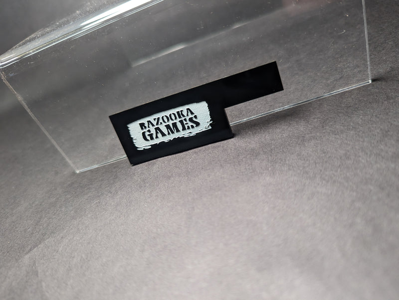 The Bazooka Games Movement Tool 3 Inch is a black rectangular tool with a silver label, shown on gray surface partly under clear acrylic. Perfect for AOS and 40k players.