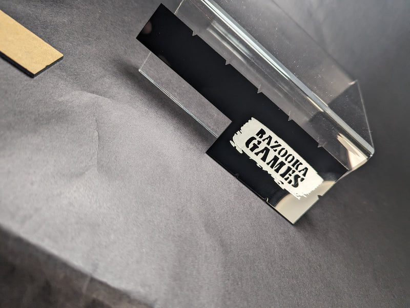A transparent plastic box with the black and white PAXORIA GAMES logo, on a dark gray surface beside brown material—perfect for storing your Bazooka Games Movement Tool 6 Inch.