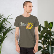 A man with short hair and tattooed forearms wears an olive green T-shirt with a cartoon bear shot from a cannon, near houseplants.