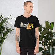 A man with short hair and tattooed arms wears a Bazooka Games T-shirt featuring a cartoon bear, standing between leafy plants.