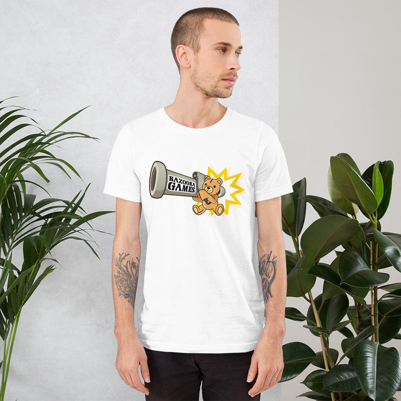 A man with short hair and tattooed arms wears a Bazooka Games T-shirt with a cartoon bear, standing by green plants indoors.